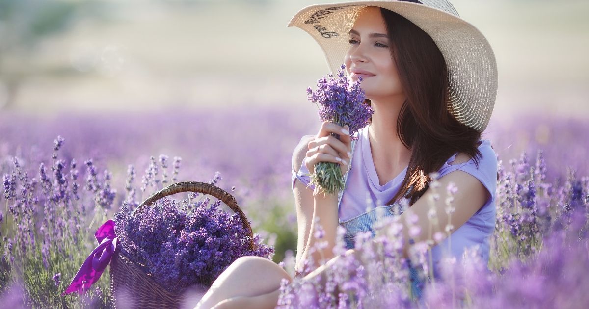 A woman wearing a hat smelling a bunch of lavender flowers in her hand. She sits in a purple lavender field. 